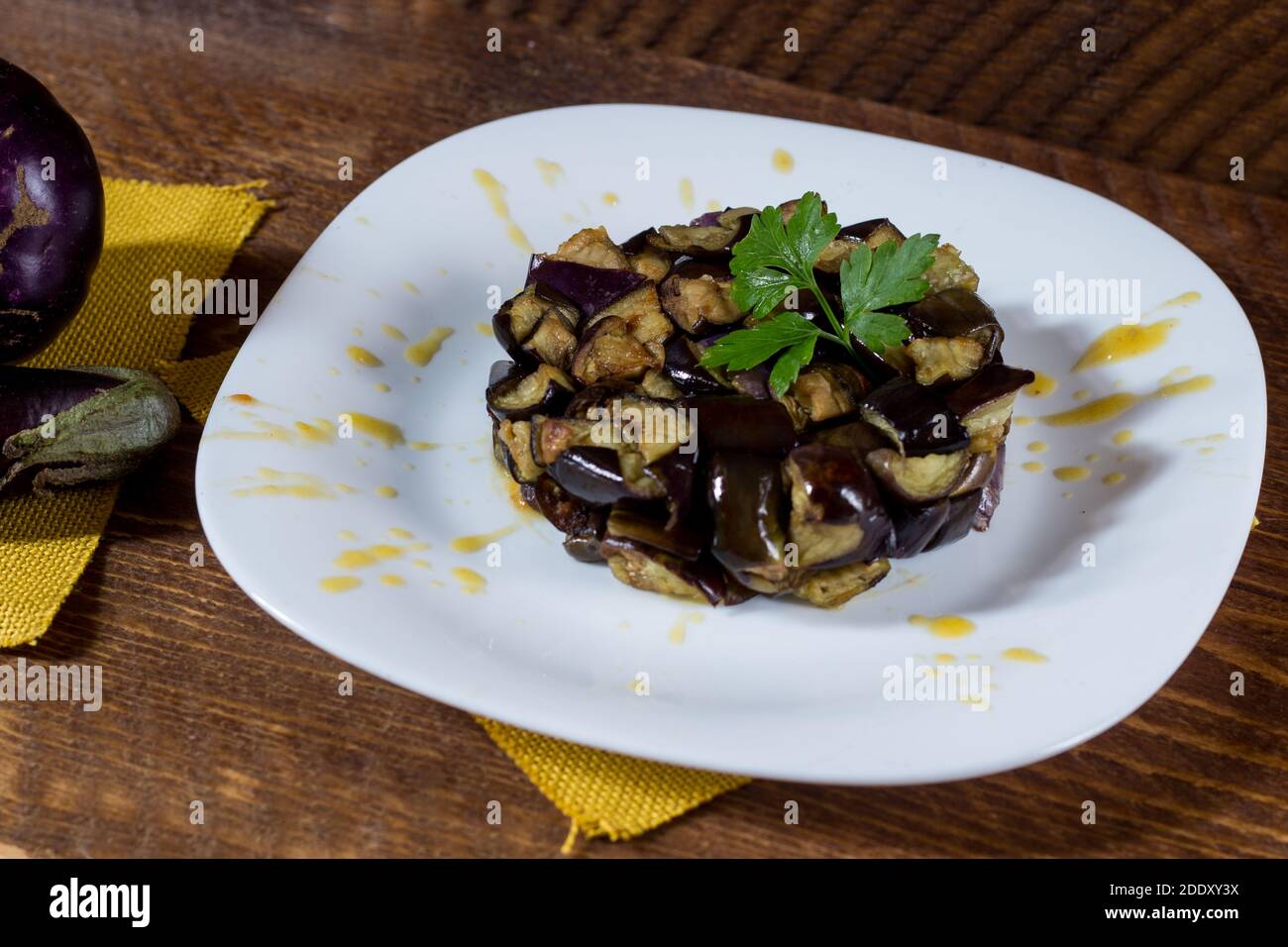 Plate of diced aubergines on a cream of yellow cherry tomatoes in a white plate. Wood background. Selective focus. Stock Photo