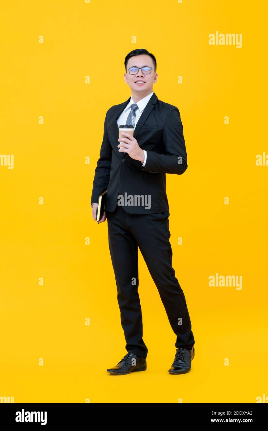 Full body studio portrait of Asian businessman in formal suit holding book and coffee cup standing in yellow background Stock Photo