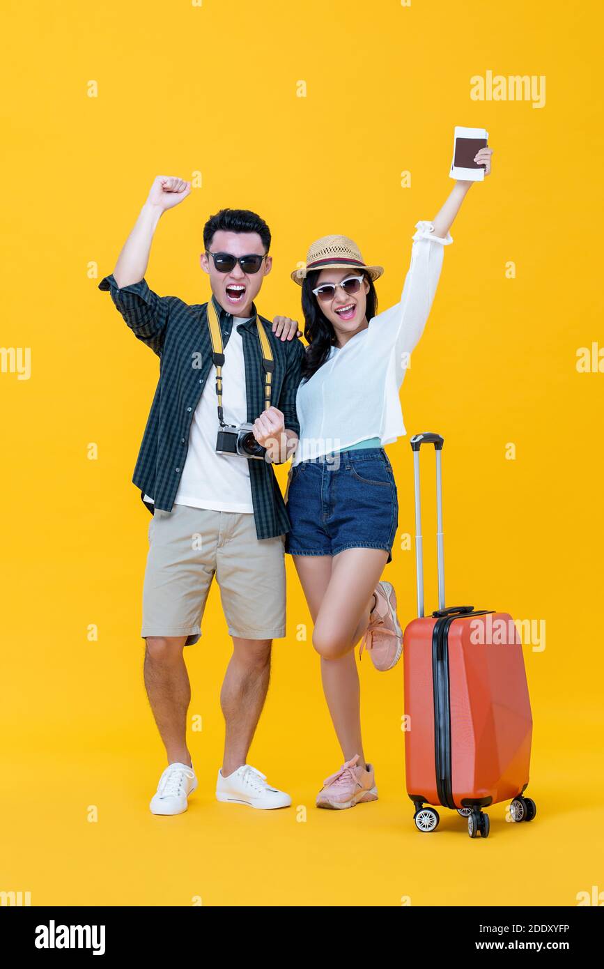 Excited Asian couple tourists with luggage raising hands up and yelling on studio yellow background Stock Photo