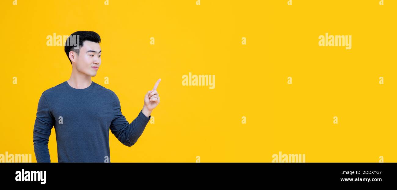 Young handsome Asian man with thoughtful expression looking sideways and pointing finger to copy space isolated on yellow banner background Stock Photo