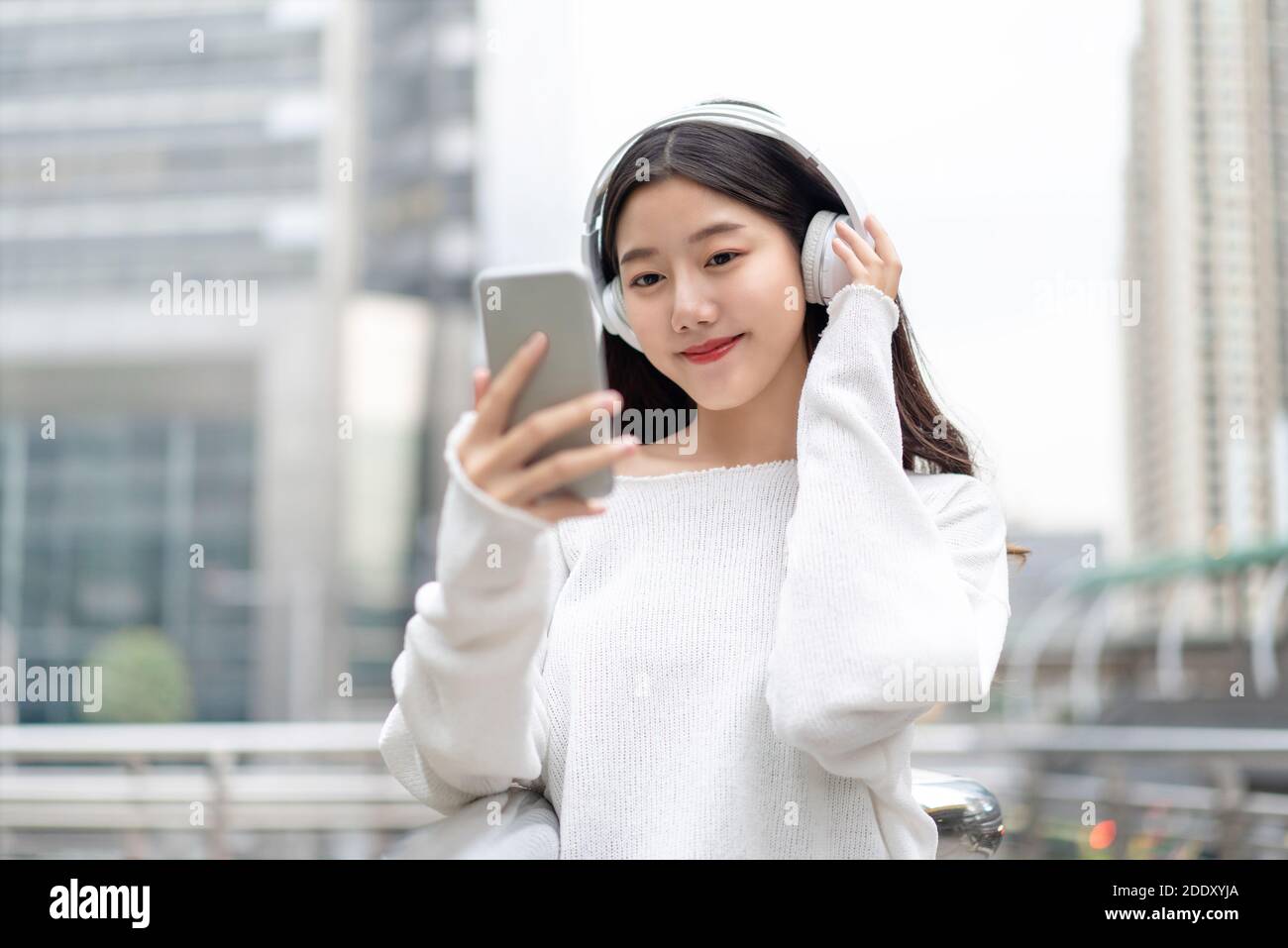 Young Asian girl wearing headphones listening to music online from mobile phone in city building backrgound Stock Photo