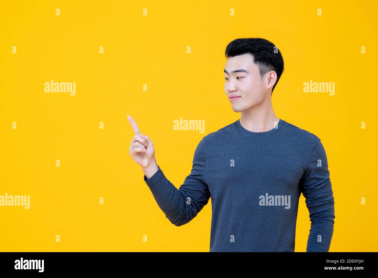 Young handsome Asian man with thoughtful expression looking sideways and pointing finger aside to empty space isolated on yellow background Stock Photo