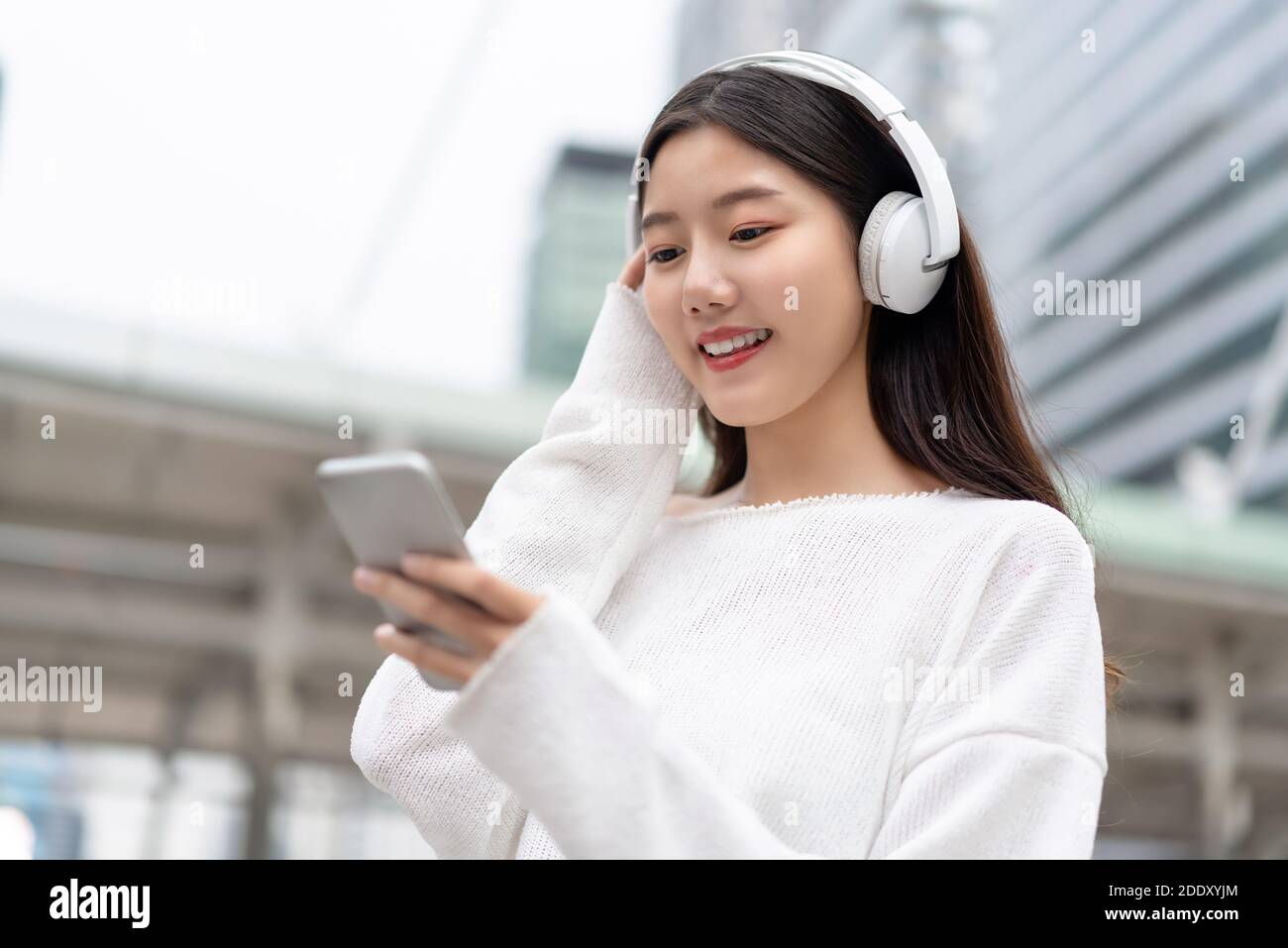 Happy smiling Asian girl wearing headphones and looking at smartphone while listening to streaming music against city building background Stock Photo