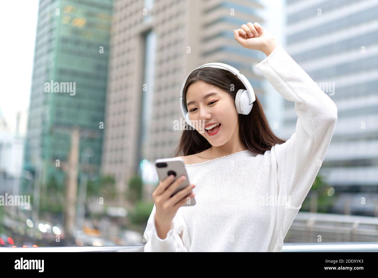 Young happy Asian girl wearing headphones listening to music online from smartphone and raising hand up in city building backrgound Stock Photo