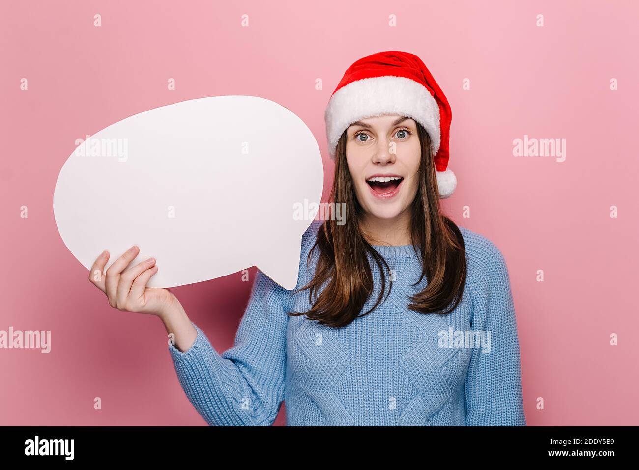Happy shocked young woman holding blank speech bubble, dressed in Christmas red hat and cozy blue sweater, models over pink studio background Stock Photo