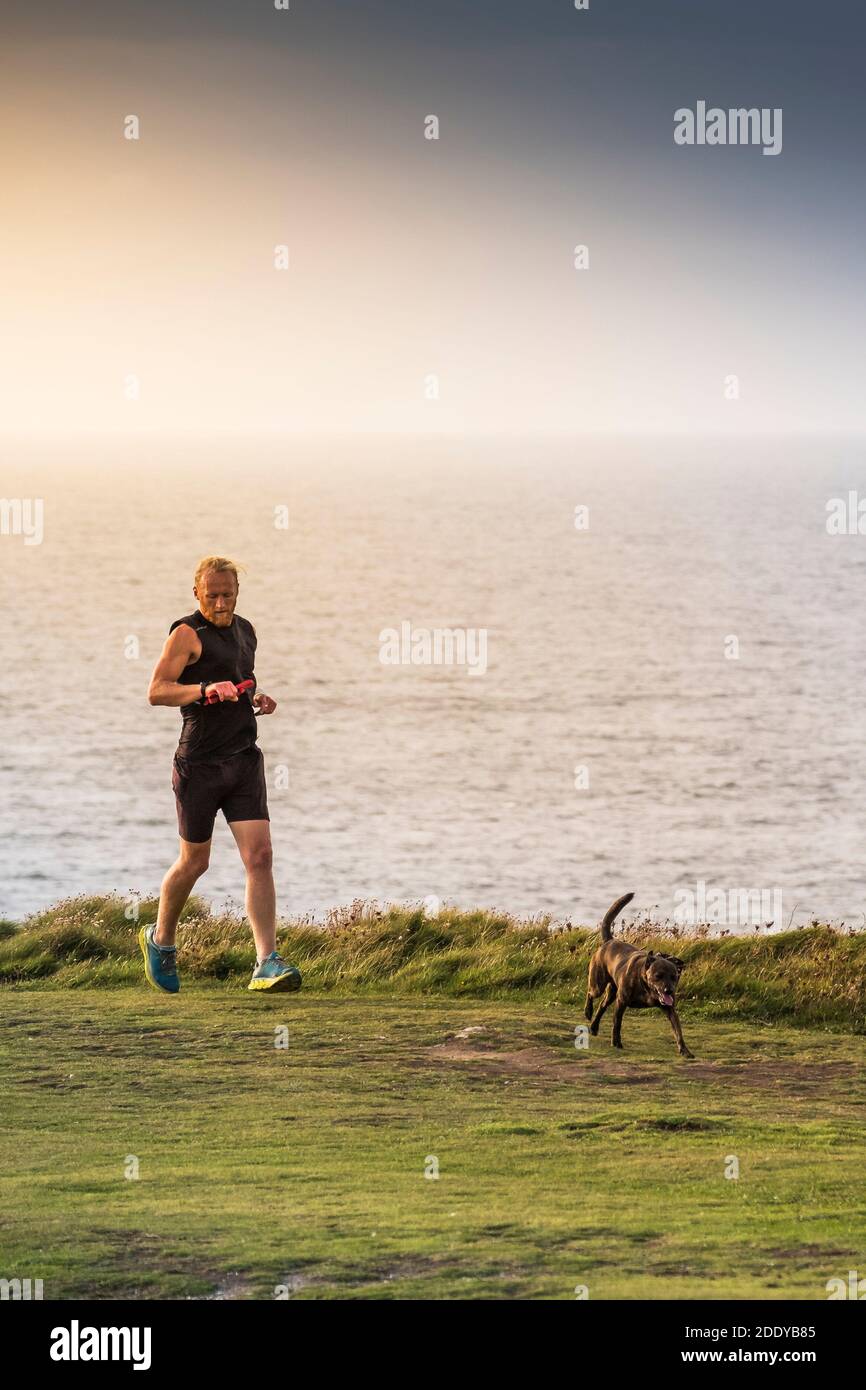 A man checking his time jogging along a footpath with his pet dog on Pentire Point East in Newquay in Cornwall. Stock Photo