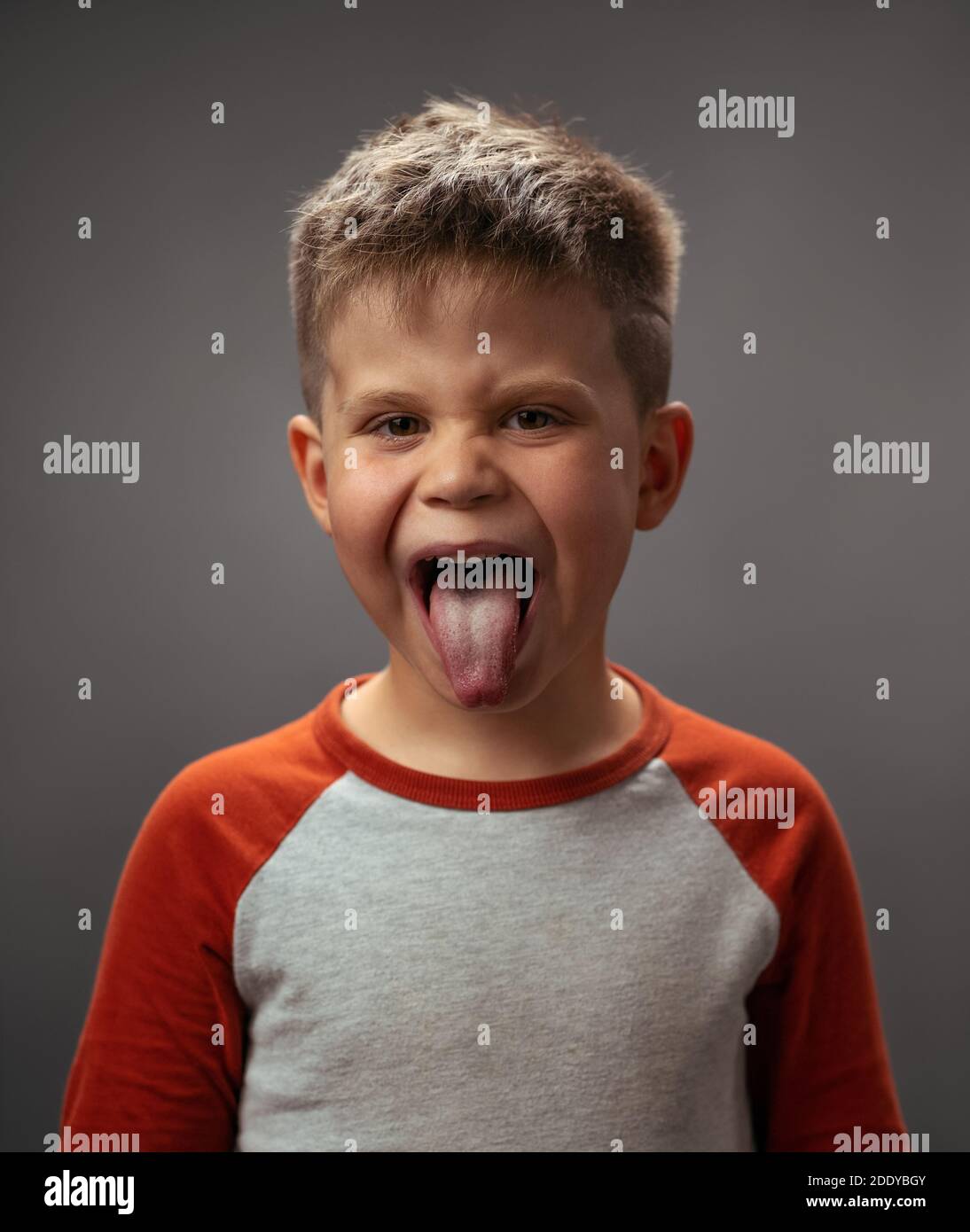 Sticking out tongue boy disobedience, bad behavior. Funny laughing boy in red shirt with eyes shut. Happy kid on dark grey background. Funny family Stock Photo