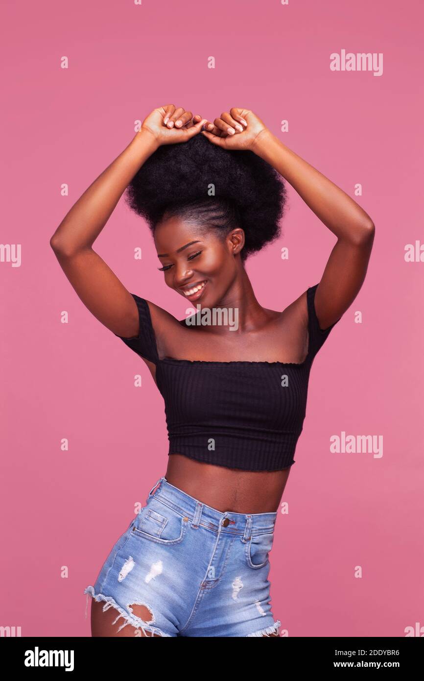 Gorgeous young African American girl with afro hair posing with arms lifted up dressed in black top and denim jeans on dirty pink background Stock Photo