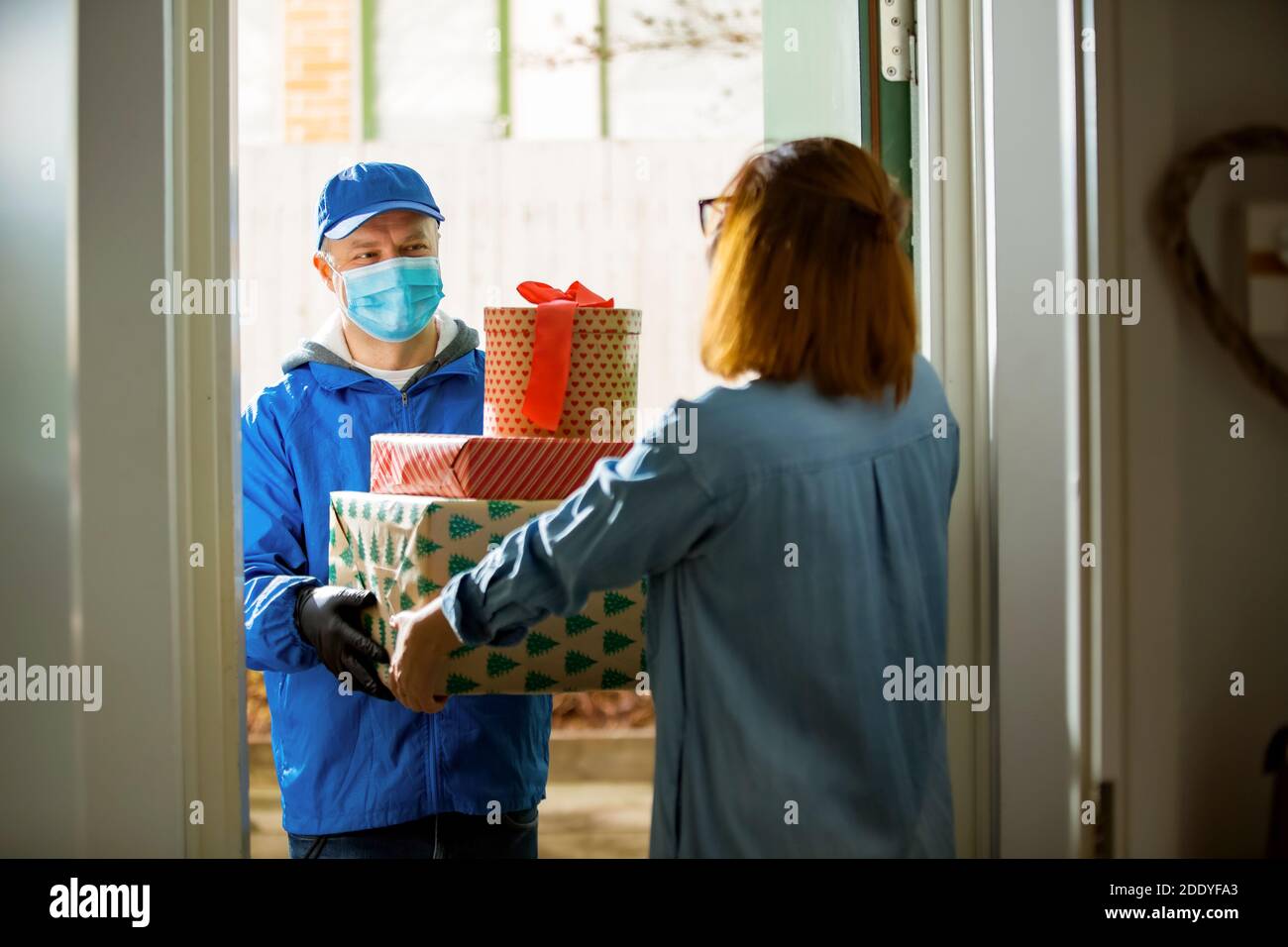 Delivery man bringing holiday packages. Woman at home standing in doorway, receiving parcels for Christmas gifts. Delivery guy in protective mask Stock Photo