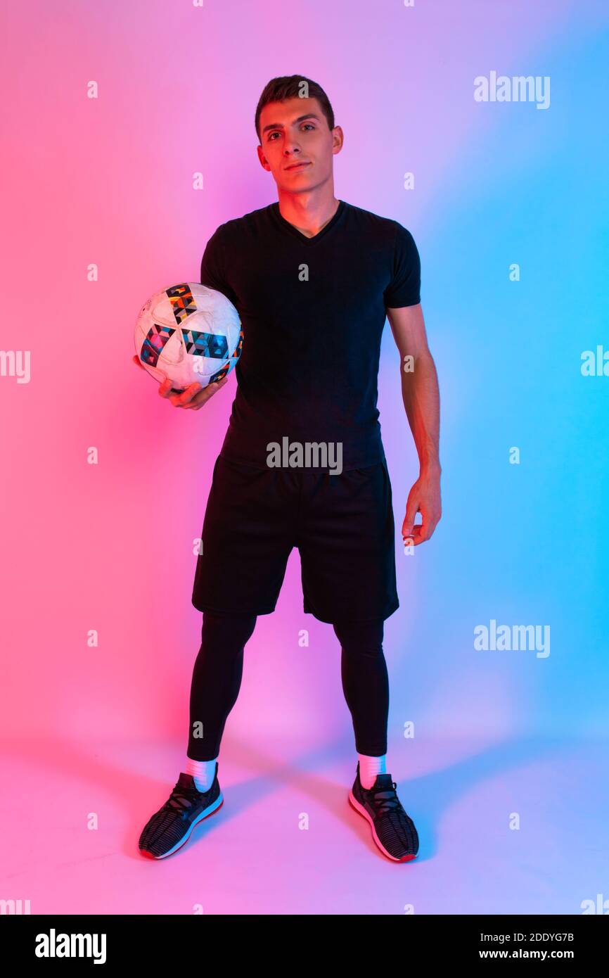 Football player in sportswear, posing holding a football, standing on a background with red and blue neon light Stock Photo