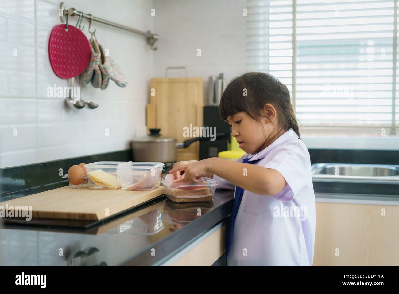 Asian elementary school student girl in uniform making sandwich for lunch box in morning school routine for day in life getting ready for school. Stock Photo