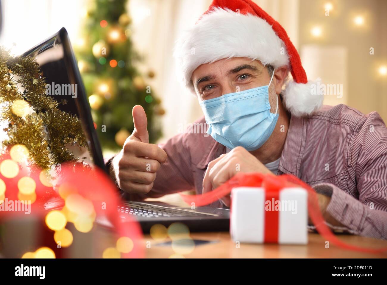 Man using a laptop at home with coronavirus mask with ok hand gesture on christmas holidays. Horizontal composition. Front view. Stock Photo