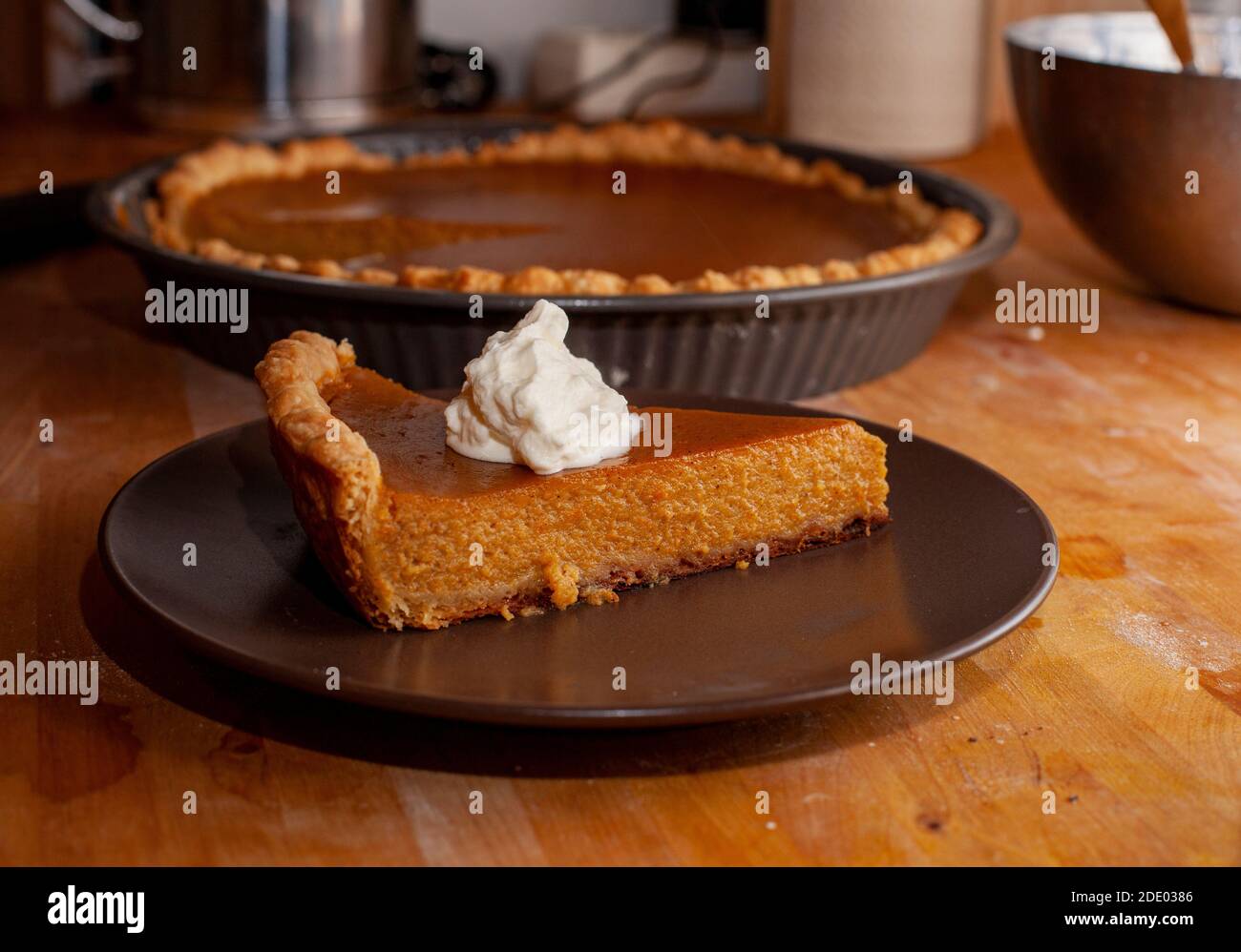 A slice of homemade pumpkin pie with a dollop of whipped cream sits on a plate on a wooden counter, with the rest of the pie in the background. Stock Photo