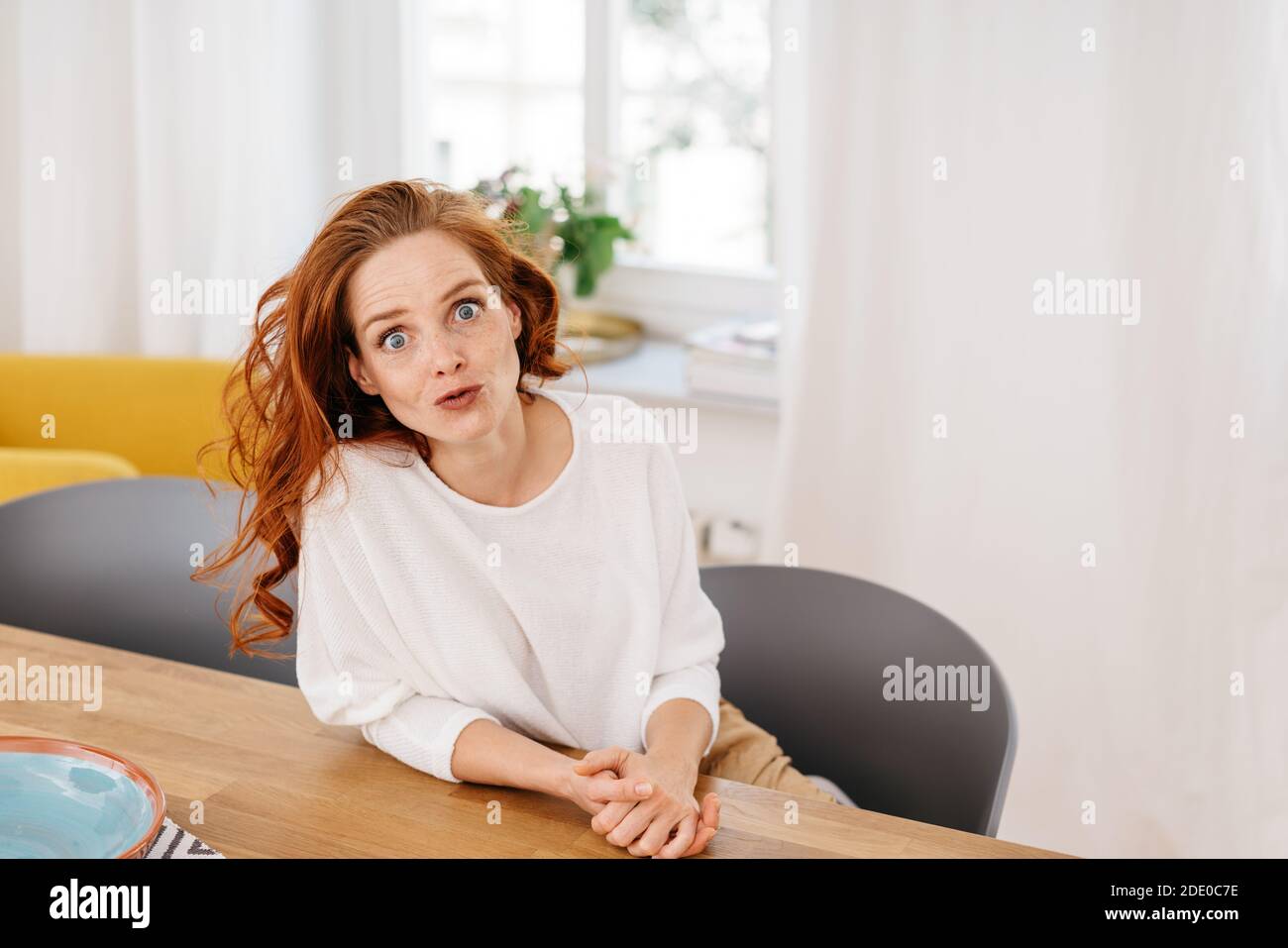 Shocked young woman staring wide eyed at the camera in disbelief or amazement as she relaxes sitting at a table at home with copyspace Stock Photo