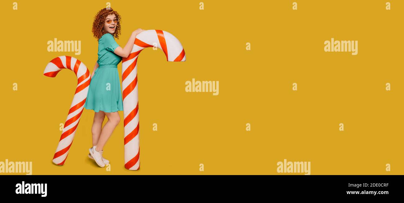 Red-haired curly woman with waw emotions stands near candy canes on fortuna gold background. Christmas and new year sweets Stock Photo
