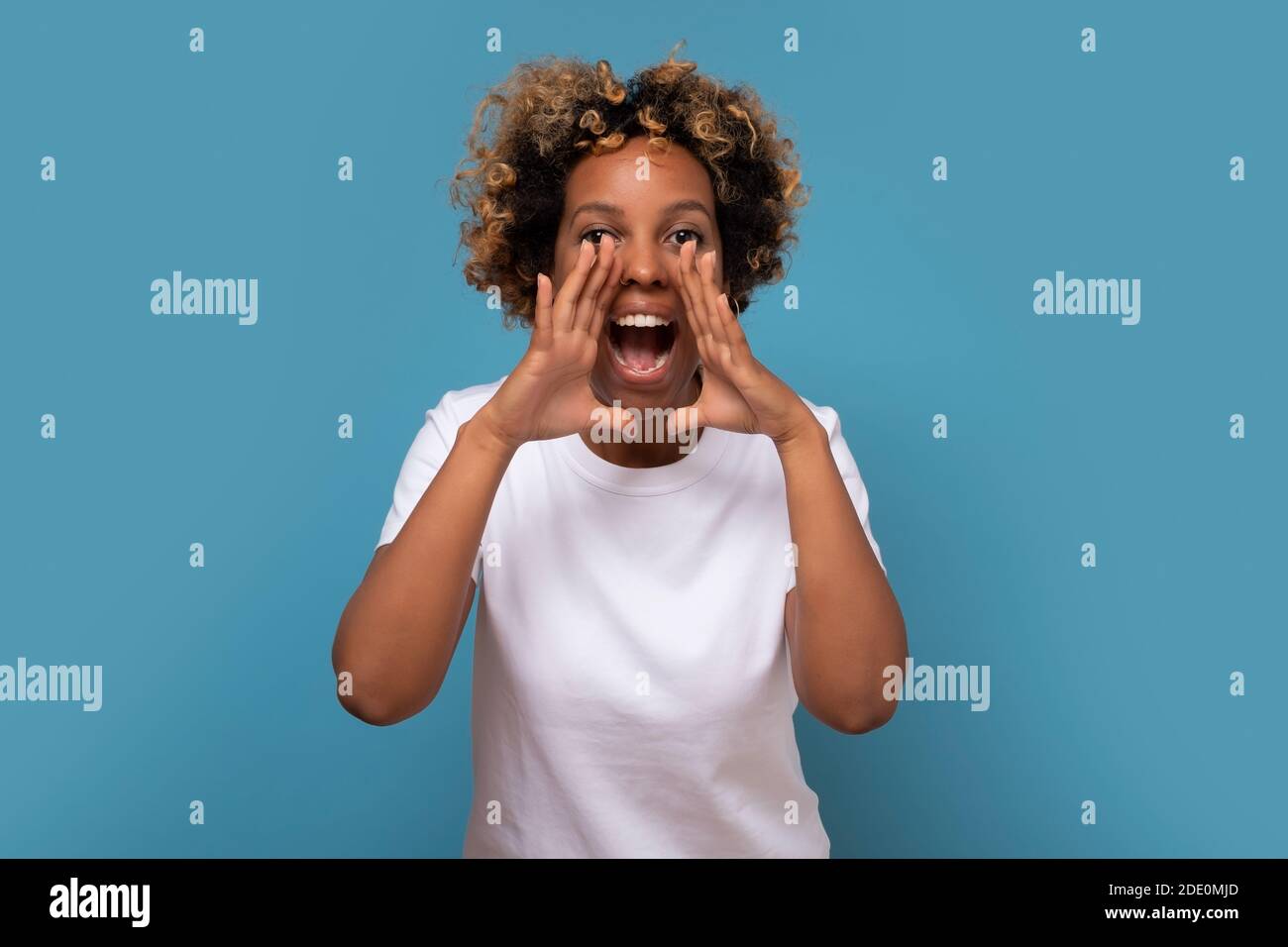 Young african woman shouting and screaming loud to camera with hand on mouth. Listen to the latest news. Studio shot on blue wall. Stock Photo