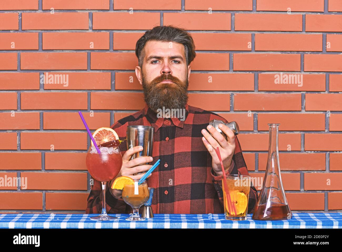 Alcoholic drinks concept. Barman with long beard and mustache and stylish hair on strict face holding shaker, makes alcoholic cocktail. Man in checkered shirt on brick wall background prepares drinks. Stock Photo