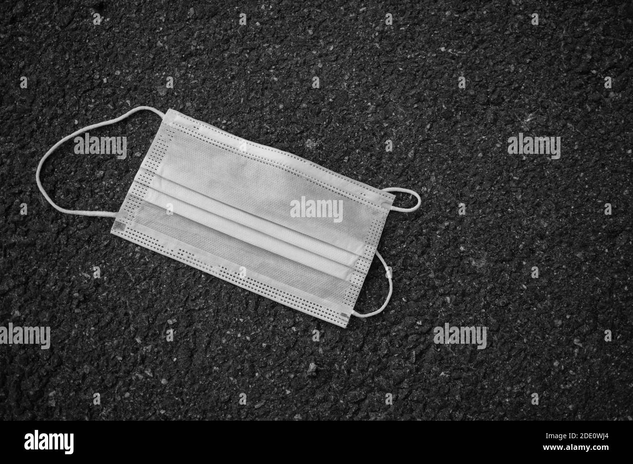 Environmental pollution during the coronavirus pandemic. White protective medical mask on dark asphalt. Black and white photo with vignetting. Stock Photo