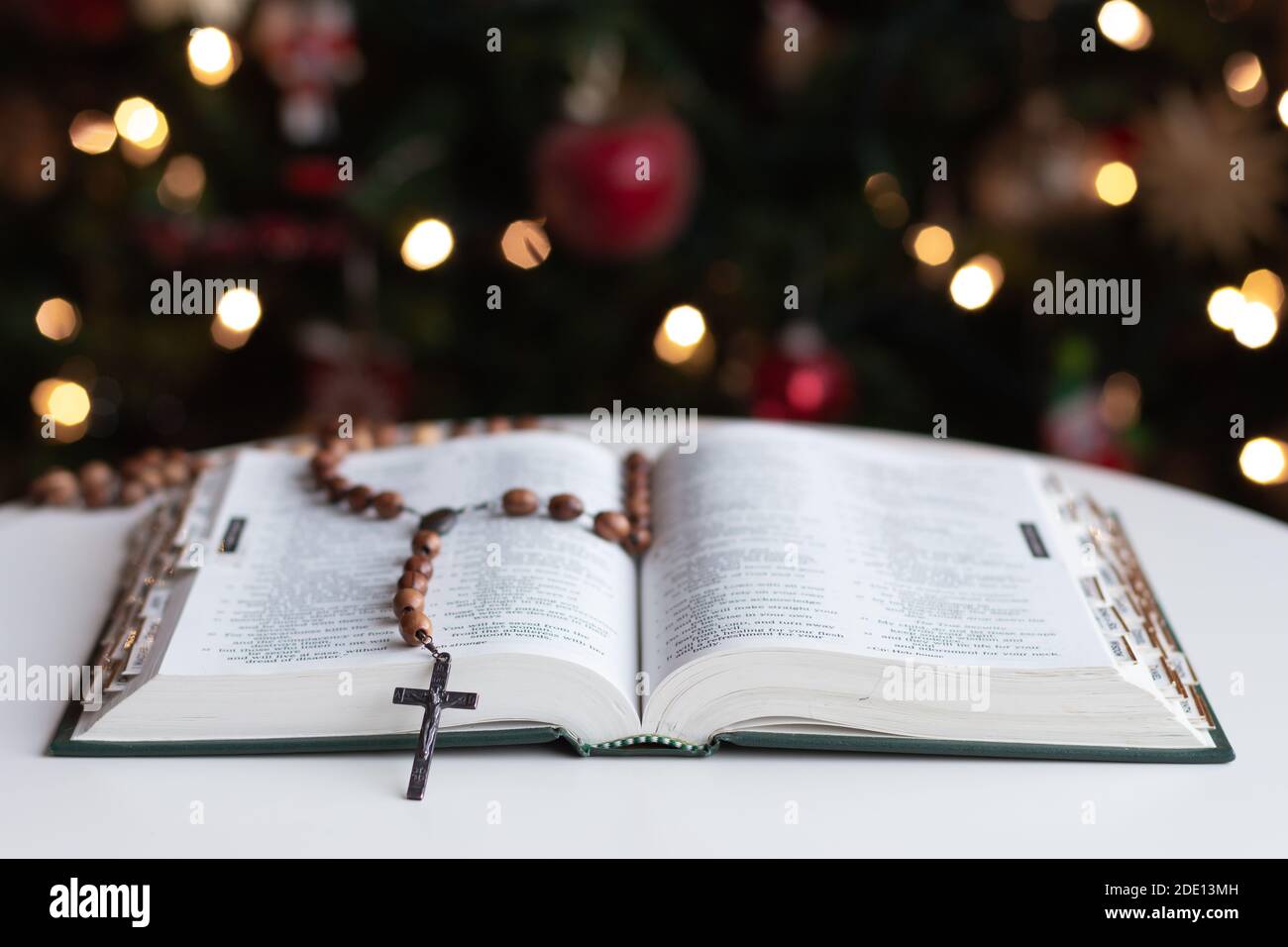Open bible with wood rosary prayer beads laying on page with Christmas tree in background Stock Photo