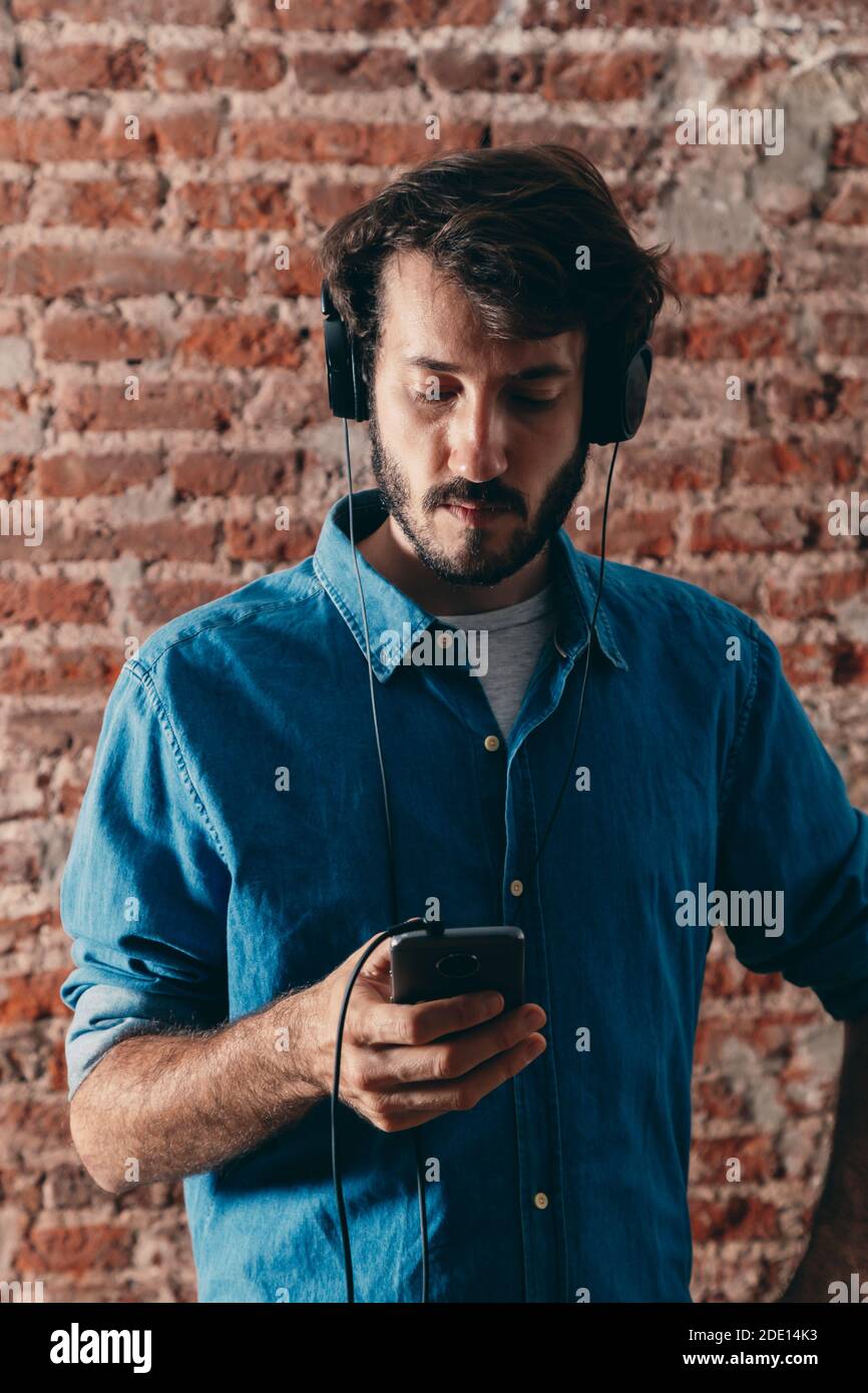 Portrait of young man with headphones and cell phone. Casual wear and brick wall of background. Stock Photo