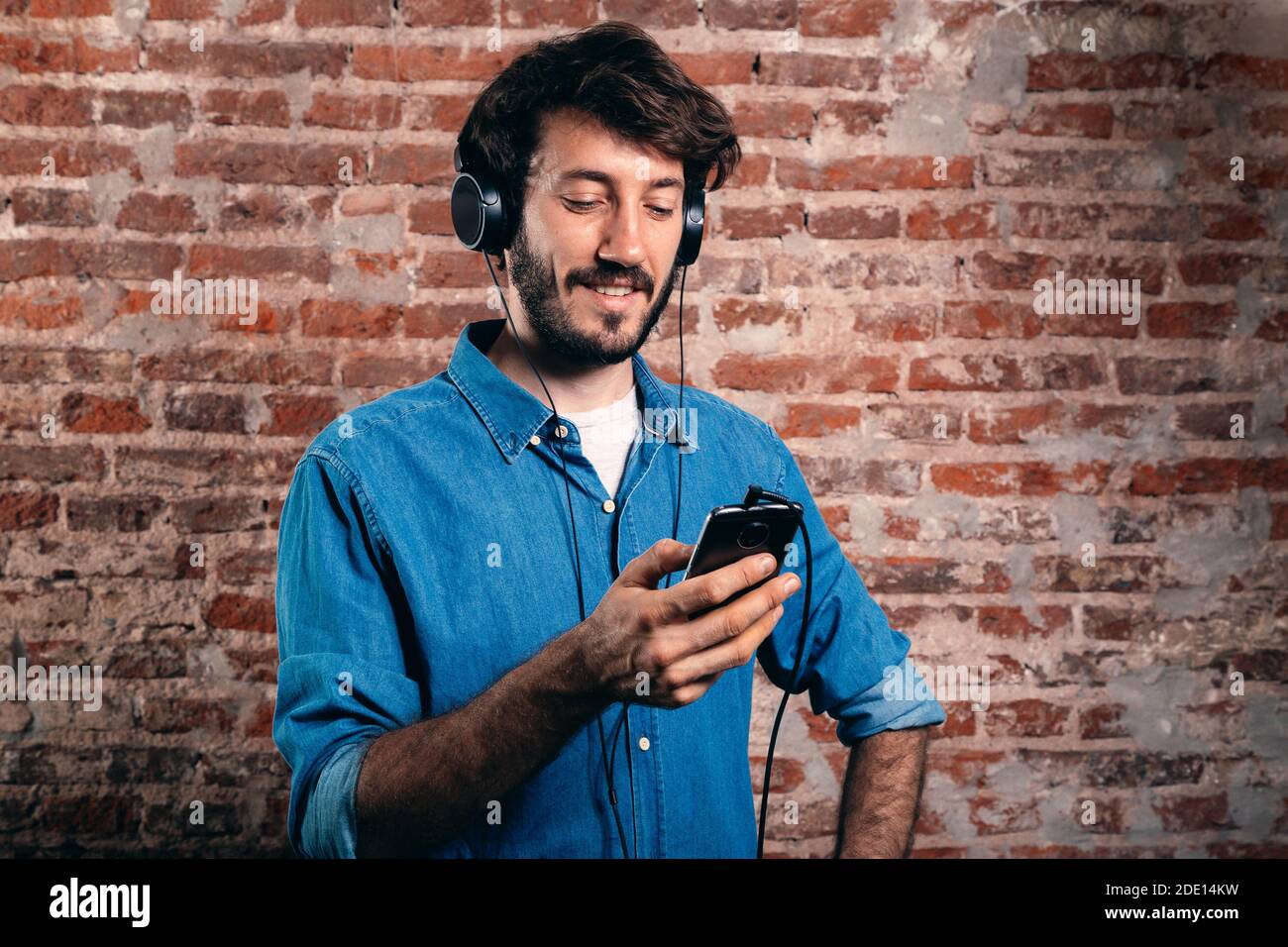 Portrait of smiling young man with earphones and cell phone. Casual wear and brick wall of background. Stock Photo