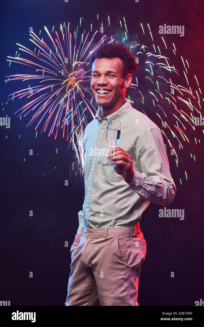 Vertical shot of a young happy mixed race man with glass of champagne standing against fireworks background, smiling at camera. New Year or Christmas party, people and holidays concept Stock Photo
