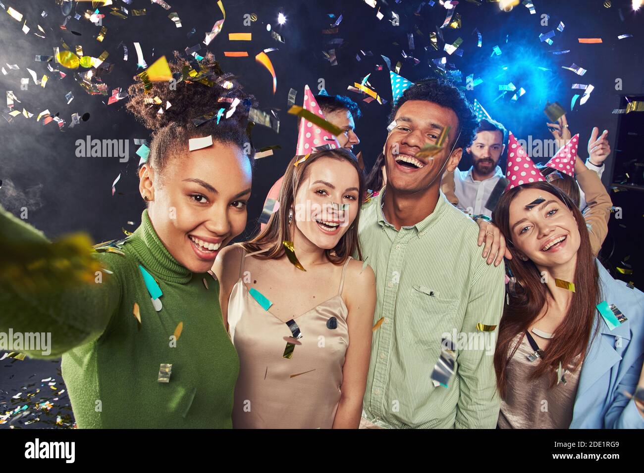 Group of young happy multiracial people wearing birthday hats making a selfie on mobile phone, confetti falling in the air. Holiday, party and celebration concept Stock Photo