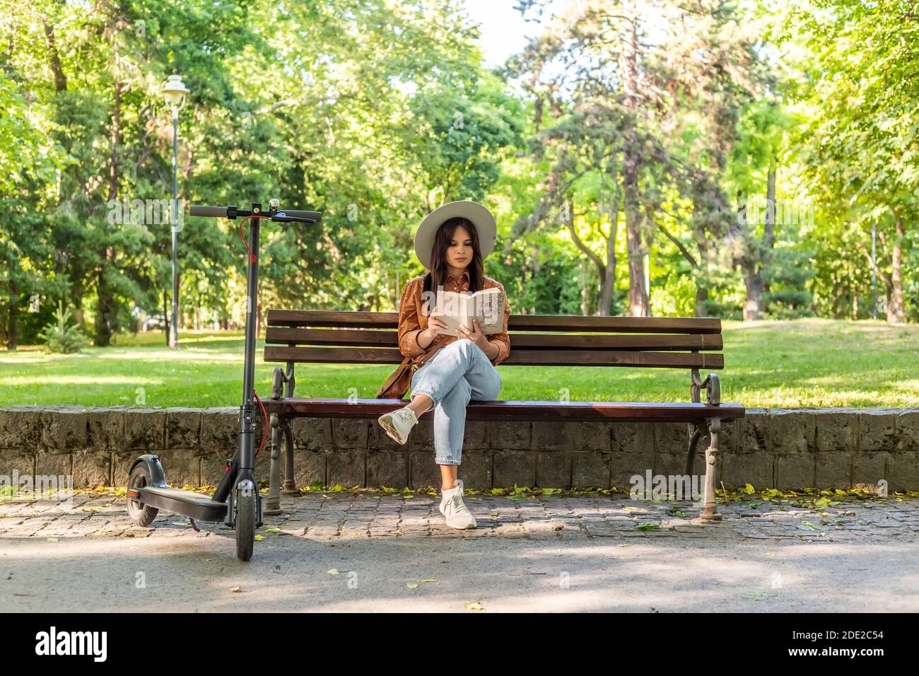 A young beautiful woman with a hat is sitting on a park bench reading a ...