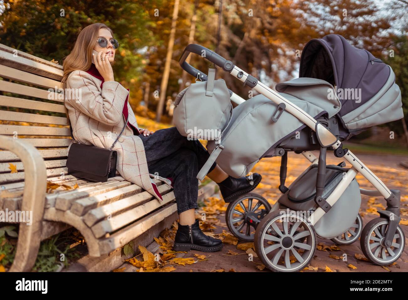 Stylish mom with stroller talking on the phone while sitting on bench ...