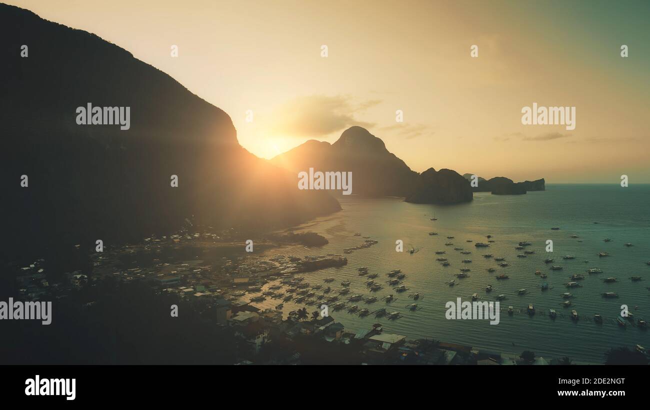 Sunset at mountainous islands at ocean harbor with dark silhouette. Amazing highlands with tropic forest and small buildings at hillside sand beach. Water transport: ships, yachts, boats at sunlight Stock Photo