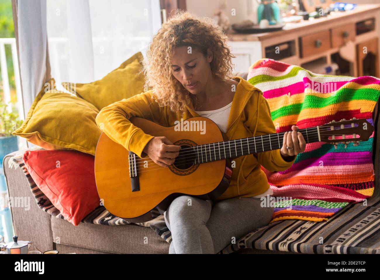 Young adult woman at home learning to play guitar - people and music concept - beautiful female playing instrument sitting on the sofa during lockdown Stock Photo