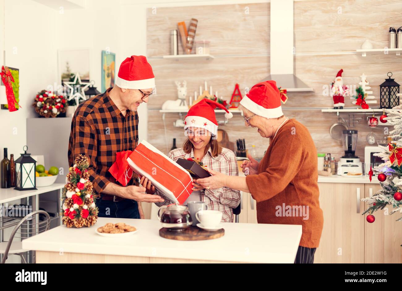 Little girl get surprised with gift boxes for chritmas from senior man and woman wearing santa hat. Happy loving grandparents celebrating winter holidays and relationship with granddaughter in home with x-mas decoration. Stock Photo