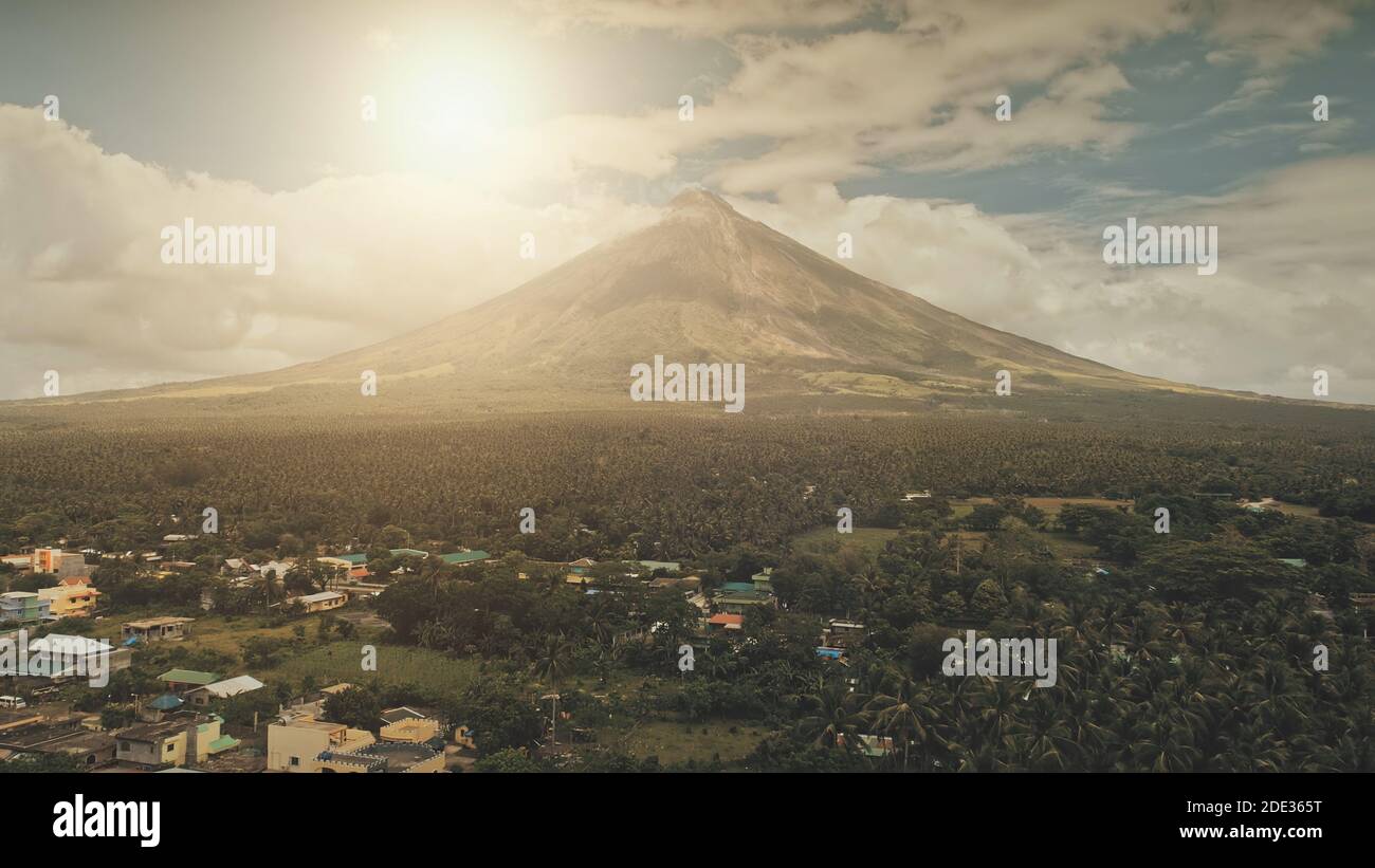 Aerial volcano erupt at sunlight closeup. Sun cityscape of rural town at green volcano valley. Contryside city streets at hillside dale of Mayon mount, Philippines. Houses, cottages at tropic palms Stock Photo