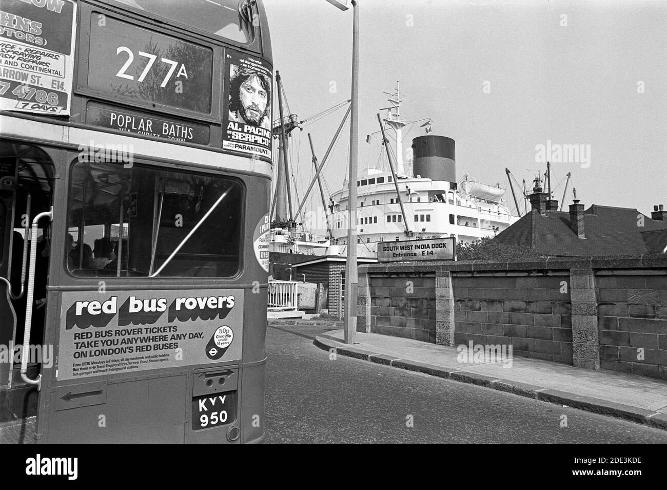 UK, London, Docklands, Isle of Dogs. West India south dock entrance, Preston's Rd / Manchester Road E14. London double-decker red bus (type RT) stopped at the raised Blue Bridge, heading north, as a ship passes through the channel that runs between the River Thames & the West India south dock. Al Pacino 'Serpico' film poster and advertising for local garage Barlow & John's Motors of Narrow Street E14. Factory employee housing over the wall to right. Stock Photo