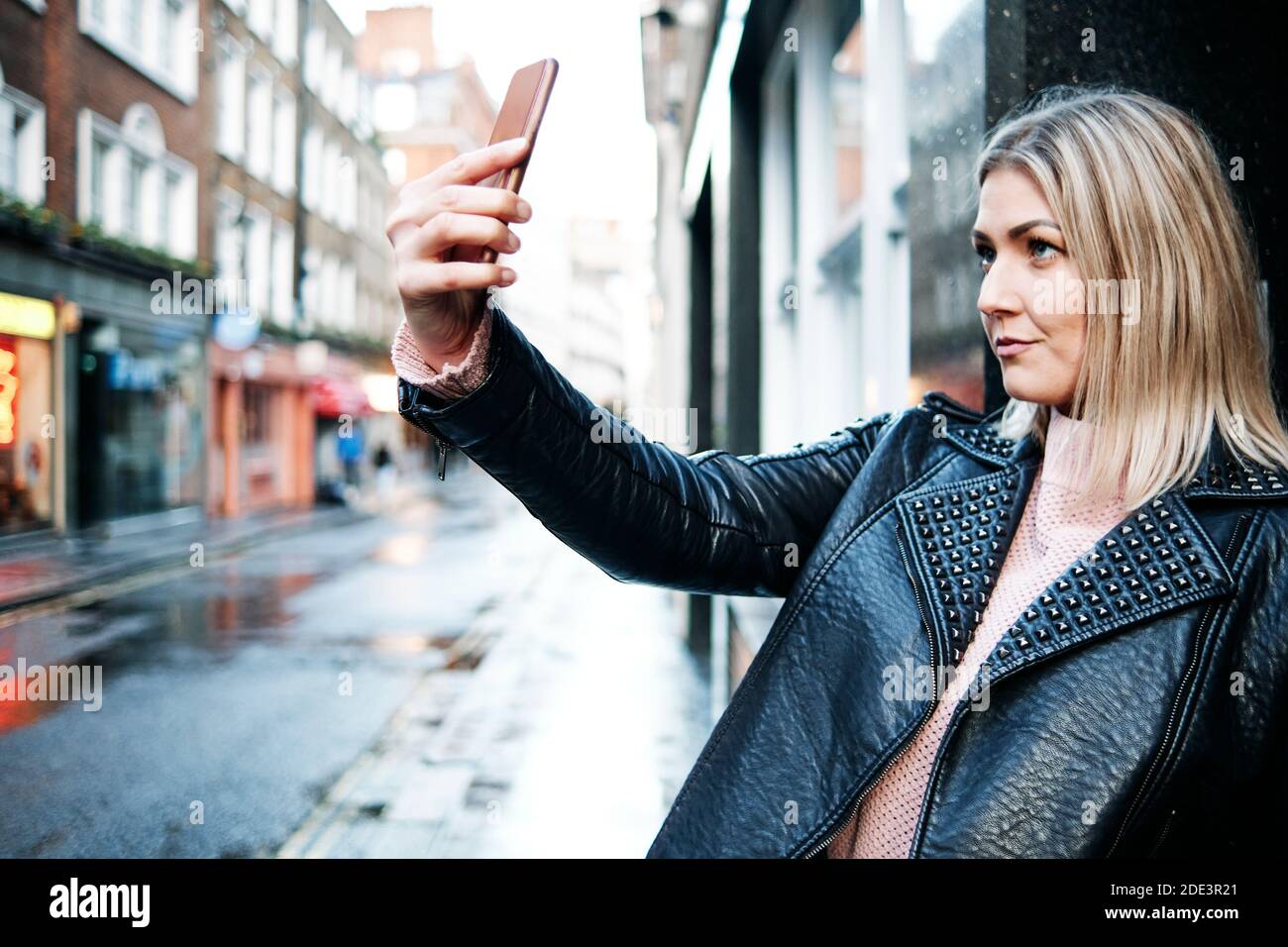Stylish Girl Biker in Fashionable Clothes. Blond Hair Blogger Woman Making Selfie on Mobile or Smart Phone. Black leather jacket. Street Fashion Lifes Stock Photo