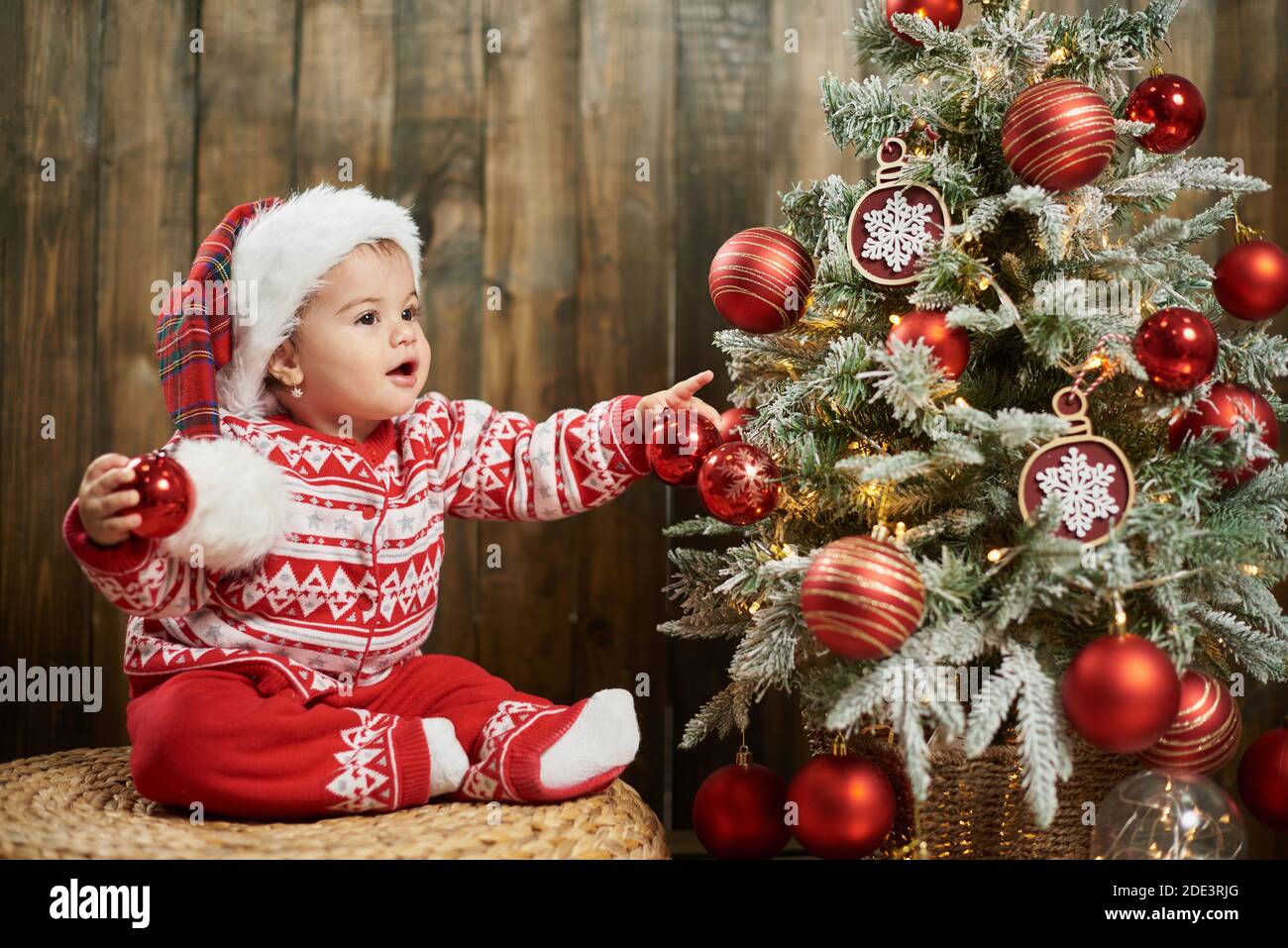 Adorable girl child wearing red sweater next to a christmas tree against wooden wall Stock Photo