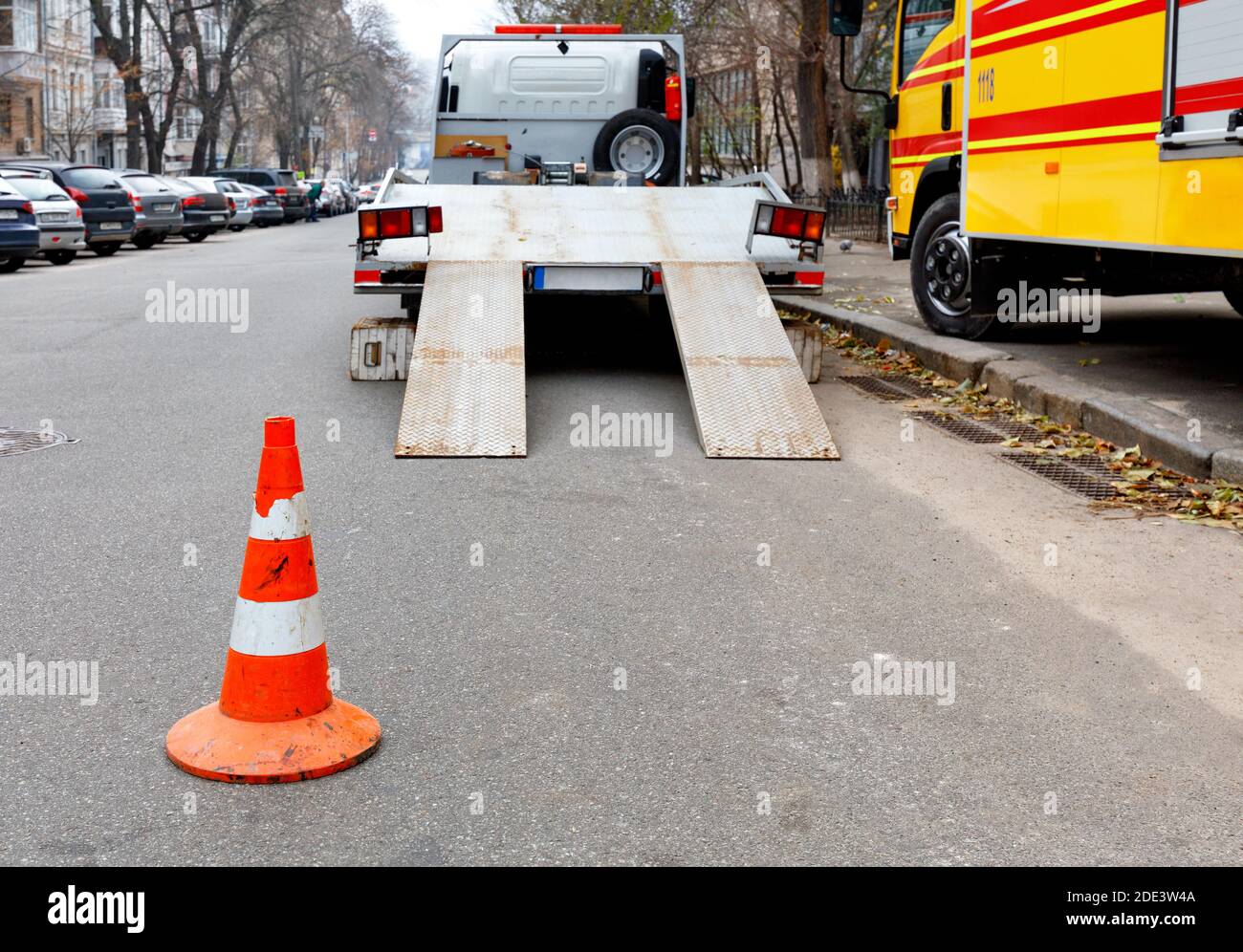 A bright orange traffic cone stands on the dark asphalt and fences the roadway with a parked transport truck. Stock Photo