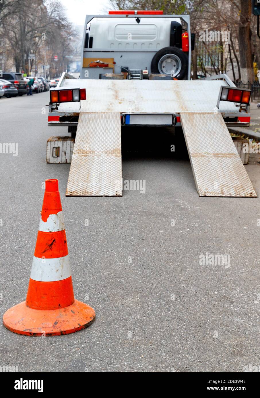A bright orange traffic cone stands on the dark asphalt and fences the roadway with a parked transport truck, vertical image, copy space. Stock Photo
