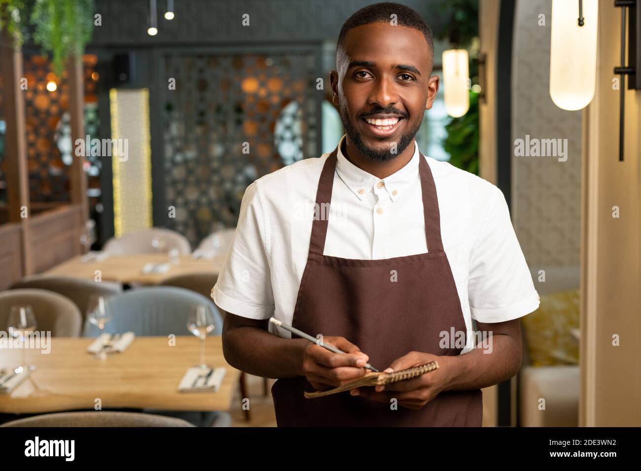 Happy young waiter with toothy smile holding pen and small notepad while standing in front of camera in restaurant and taking orders Stock Photo