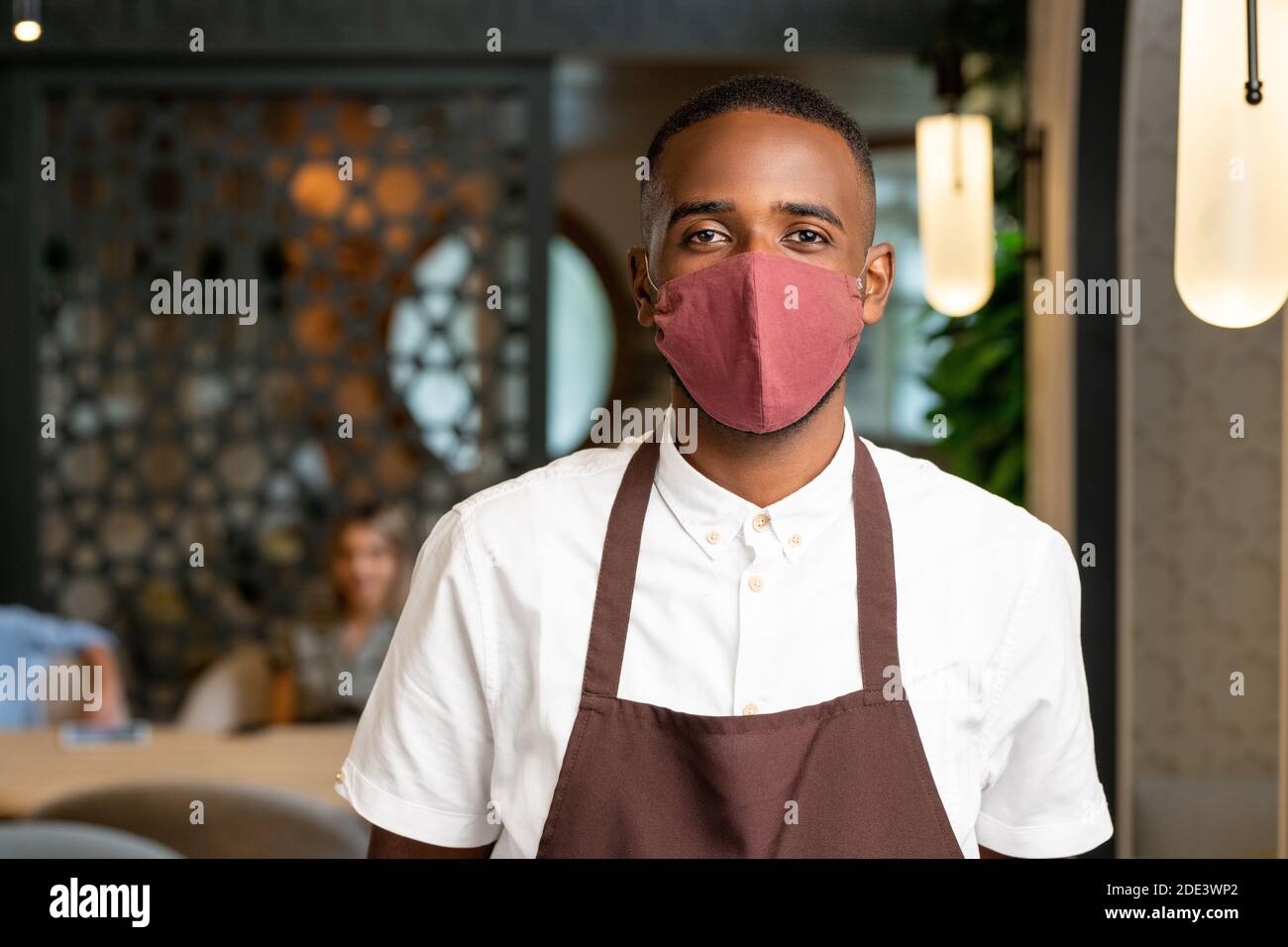 Young waiter of African ethnicity in brown apron and protective mask standing in front of camera against clients in cafe environment Stock Photo