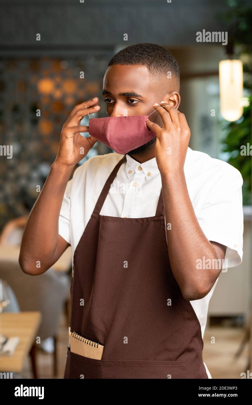 Young African man in brown apron putting protective mask on his face while going to serve new clients of restaurant where he works Stock Photo