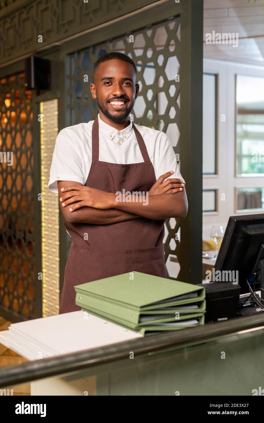 Young cheerful waiter of African ethnicity looking at you with toothy smile and crossing arms on chest while standing in restaurant environment Stock Photo