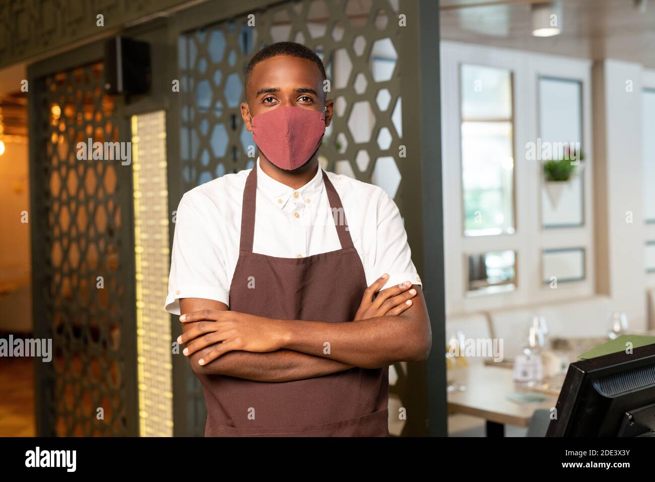 Contemporary waiter in protective mask and brown apron crossing arms on chest while standing by computer in modern cafe or restaurant Stock Photo