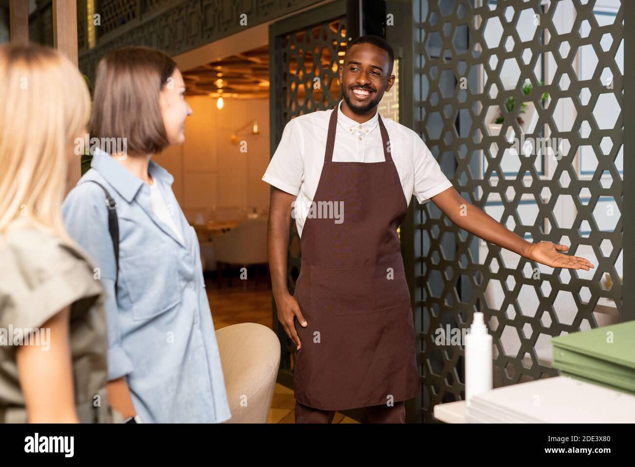 Young smiling waiter in workwear inviting two girls to one of luxurious halls of modern restaurant while looking at them and pointing at bars Stock Photo
