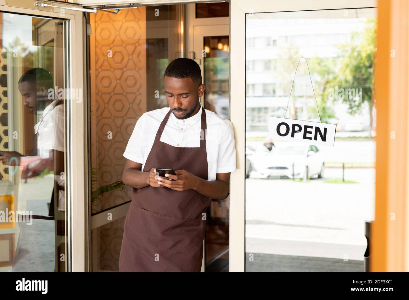 African young waiter in uniform standing in the door of cafe or resaurant and scrolling through new orders of clients in smartphone Stock Photo