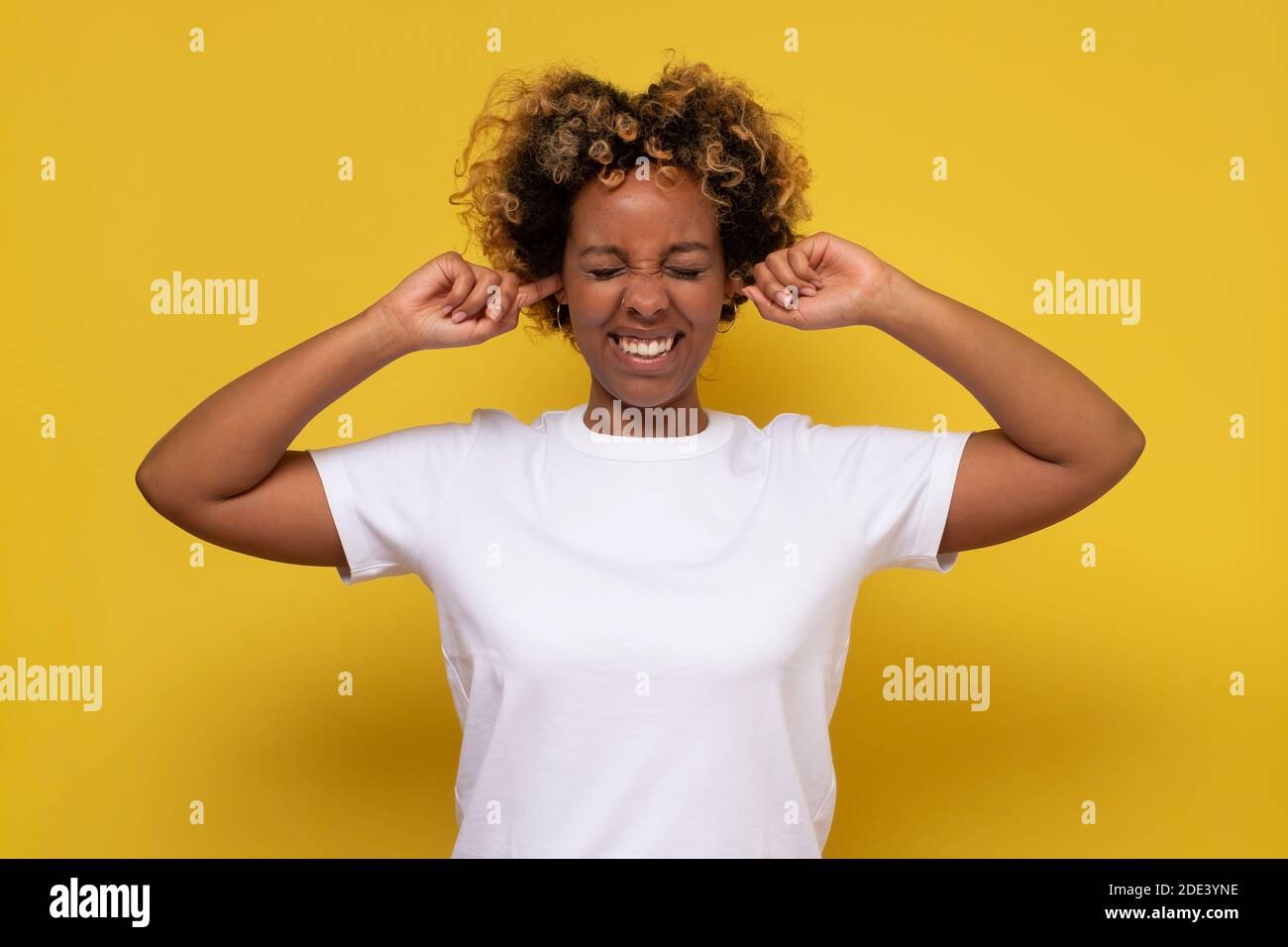 African young woman with curly hair plugging ears. Studio shot on yellow wall. Stock Photo