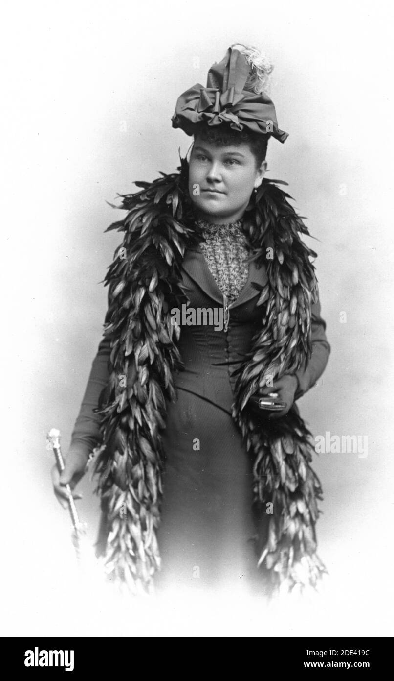 About 1890, this young woman visited her local photography studio to make a permanent image of herself wearing her fine new feather boa., which reached down to her knees. She also wears a fine hat of extravagantly wrapped ribbons topped by white feathers. She seems to be wearing no makeup. In her right hand is a silver-topped cane. She wears dark gloves, and in her left hand is a small compact, or cigarette holder, or ???  To see my other noteworthy images of interesting women, search:  prestor vintage  woman Stock Photo
