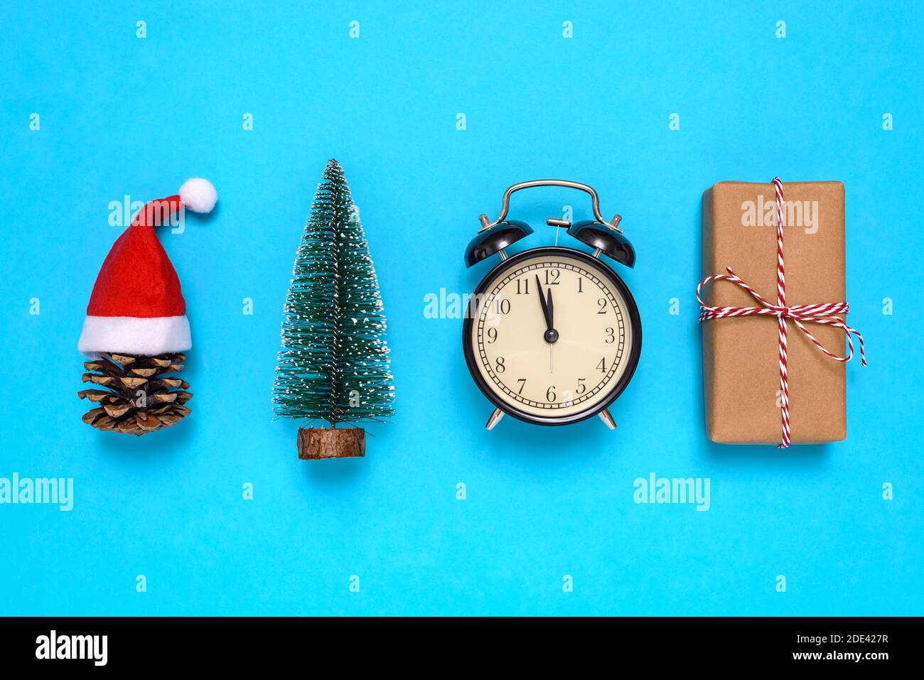 Pine cone with Santa Claus hat, small Christmas tree, alarm clock and gift box on blue background. The concept of celebrating the New Year and Christm Stock Photo