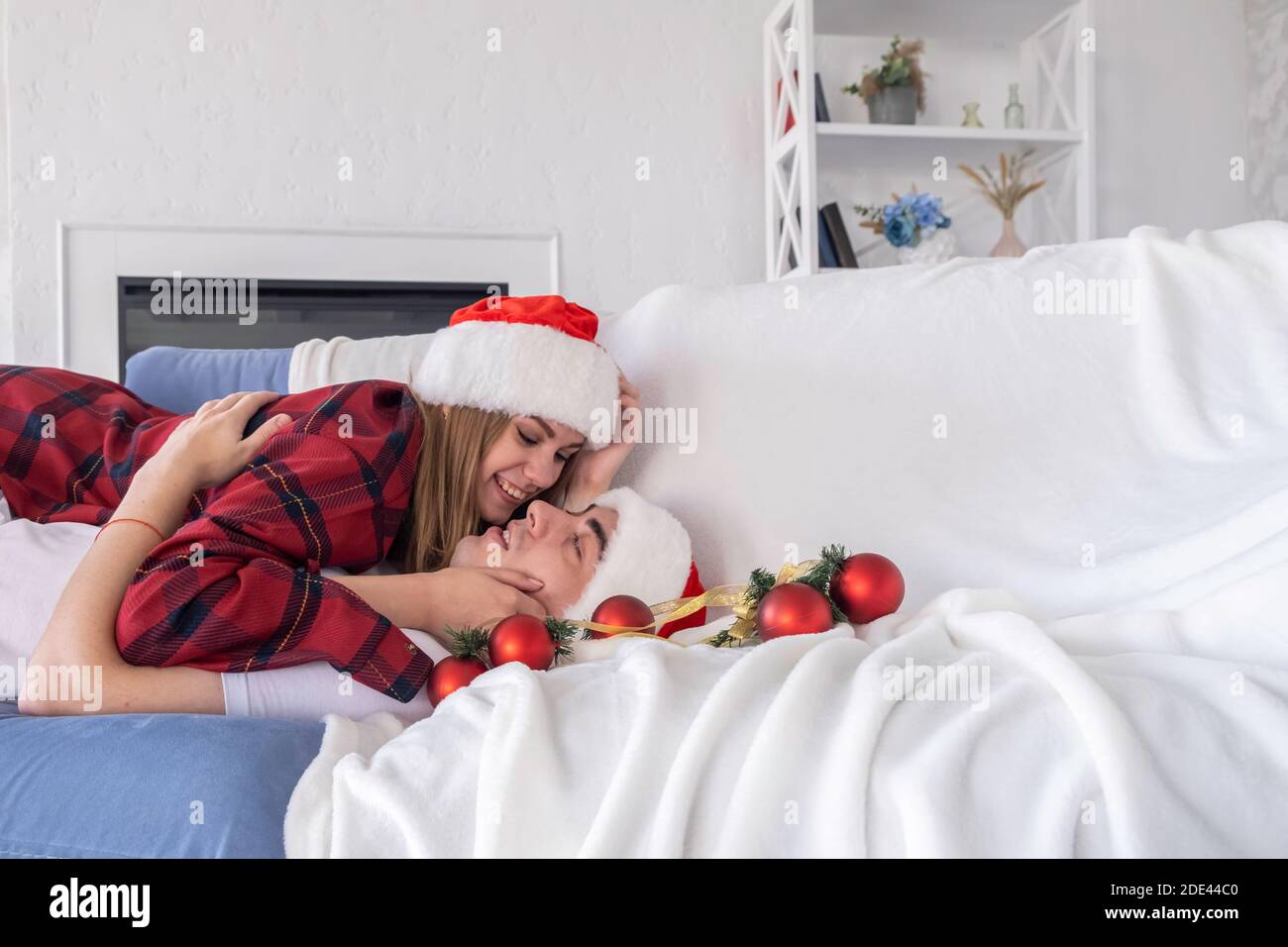 Side view of happy couple lying on couch or sofa wearing santa hats and pajamas. Young man and woman cuddling and looking at each other with tendernes Stock Photo