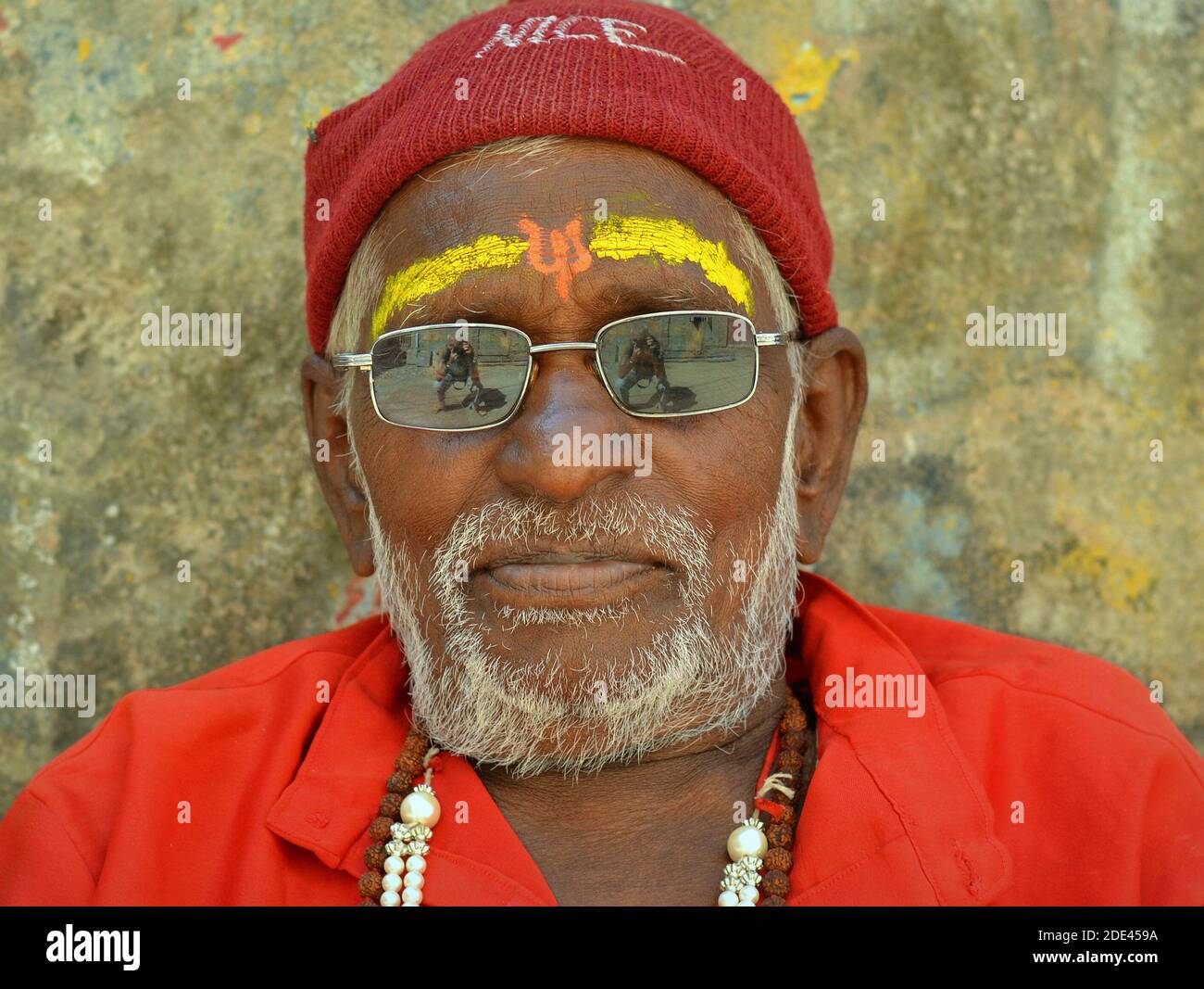 Elderly Indian Hindu pilgrim sadhu with yellow sandalwood paste and psi on his forehead wears mirrored sunglasses in front of stone wall background. Stock Photo